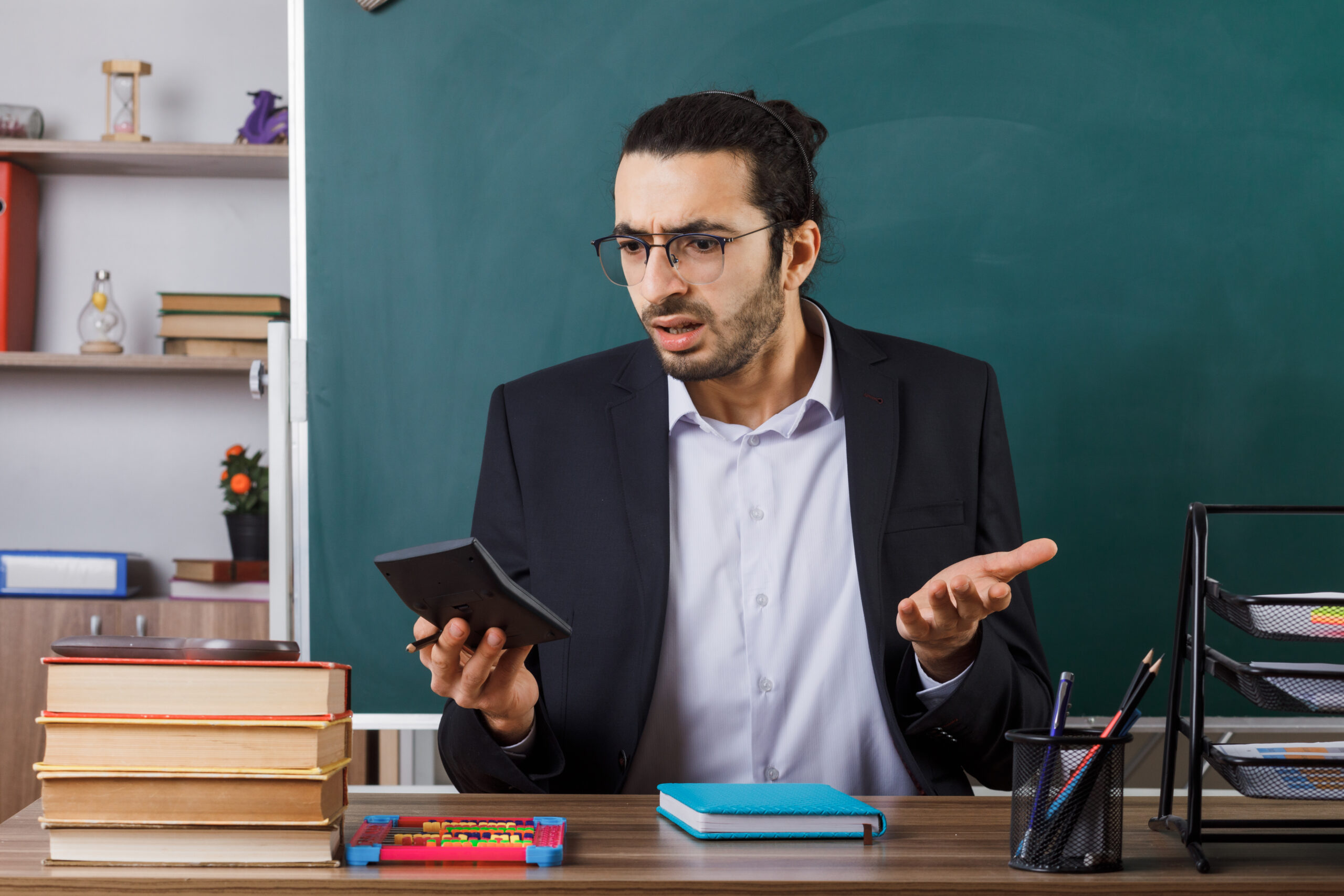 confused male teacher wearing glasses holding and looking at calculator sitting at table with school tools in classroom